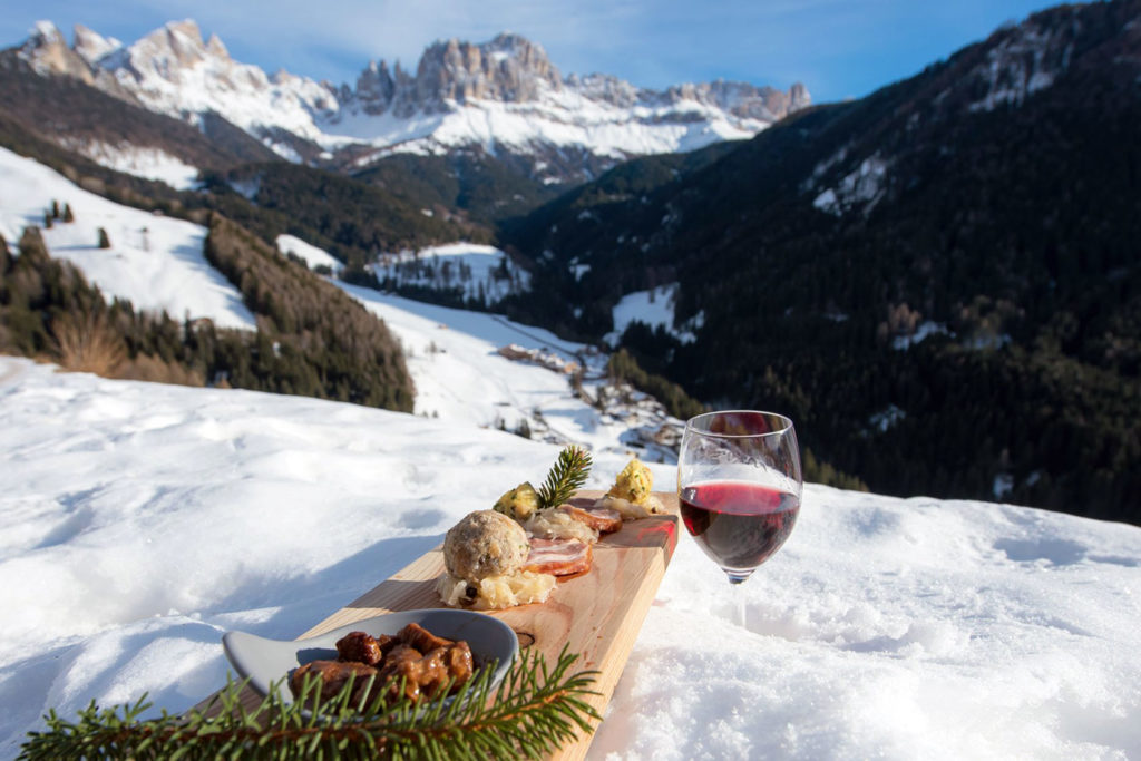 A glass of red wine in the snow next to a wooden board full of food, with a view of the snowy landscape in the distance 