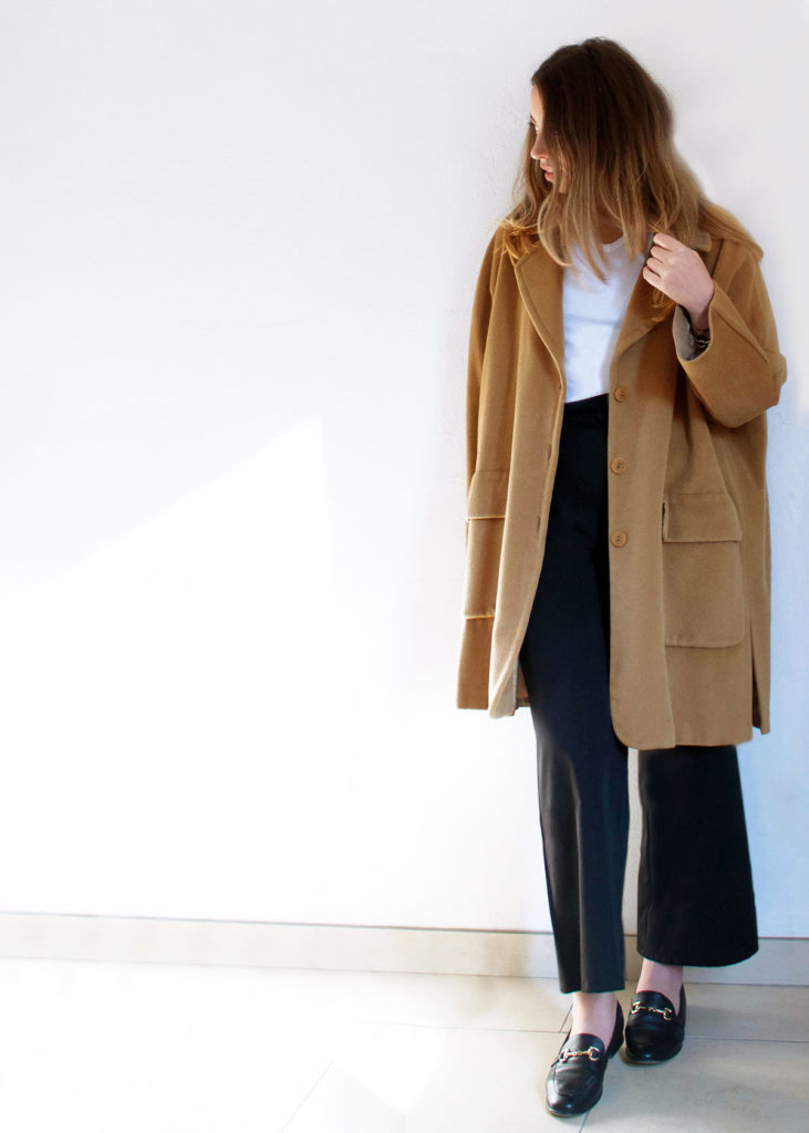 woman models long camel coat with a white T shirt, black trousers and black loafers 