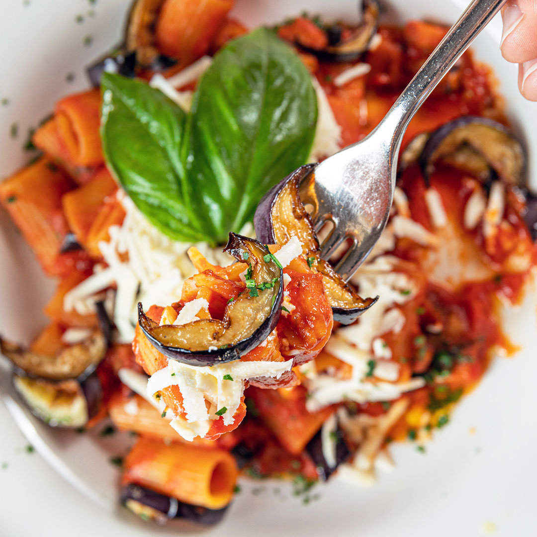 A fork taking a bite of a pasta dish with eggplants and tomatoes. 