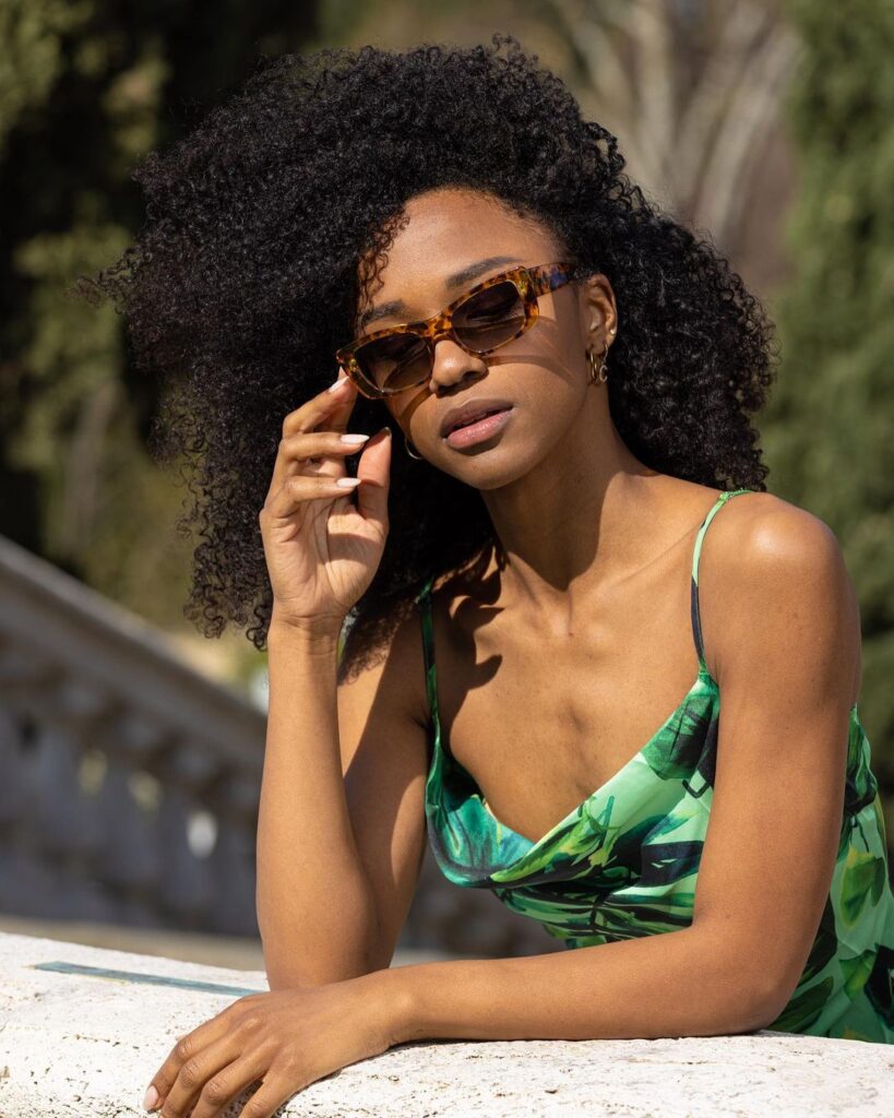 A black women models tortoise shell sunglasses and a spaghetti-strap green dress, leaning on a stone rail outside 