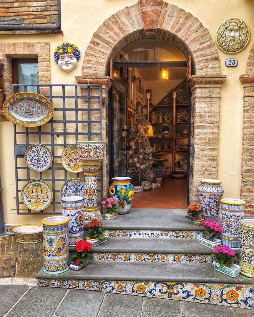 The outside of a ceramics shop in Italy. Ceramic plates and vases sit on steps in front of an arched doorway. 