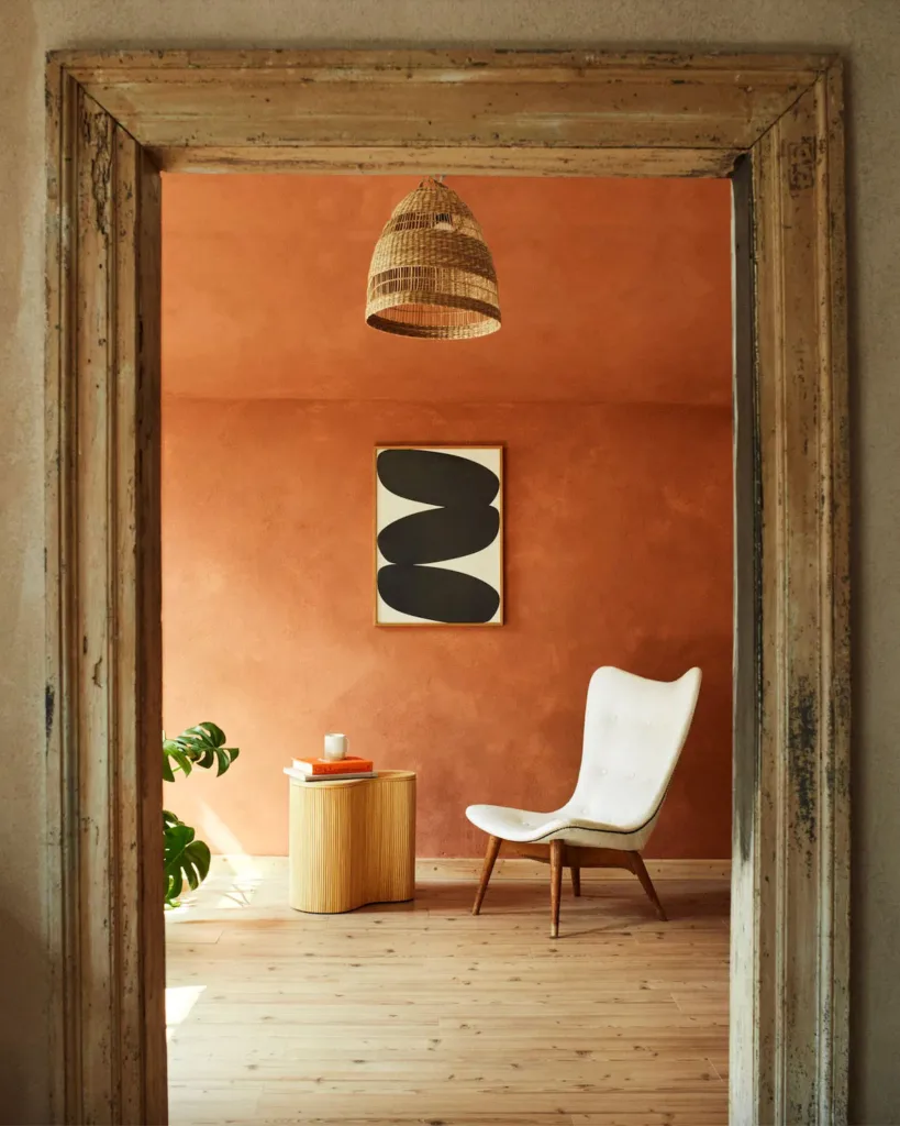 Looking into a room with orange-brown lime washed walls and wooden floor. A modern white chair sits in a corner while a woven basket light hangs from the ceiling and a white canvas with 3 thick black strokes hangs on the wall. 